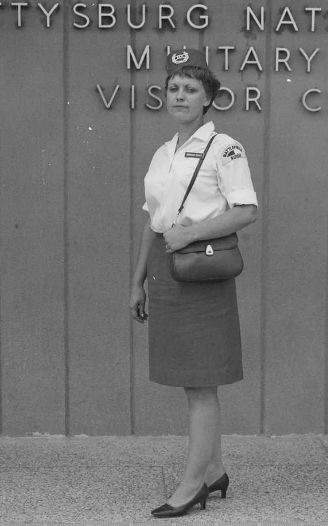 Barbara Schutt in her guide uniform with hat and purse standing in from of a visitor center.