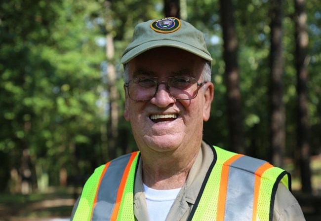 An older white man smiles in a volunteer hat and yellow safety vest.