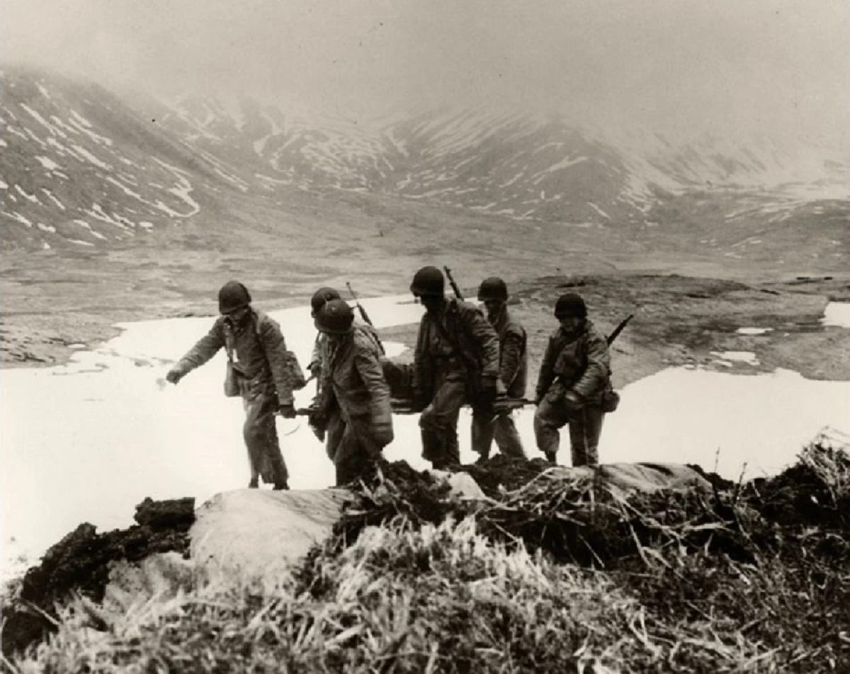 Battle of Attu 1943 black and white photo of six soldiers carrying wounded person on stretcher.