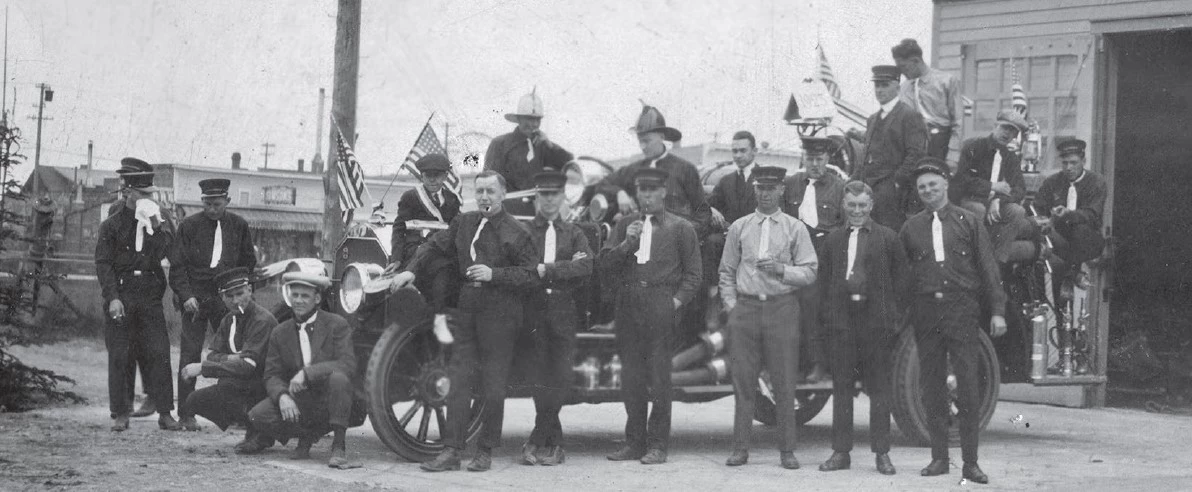 a historic scene of uniformed men standing in front of a fire engine and firehouse.