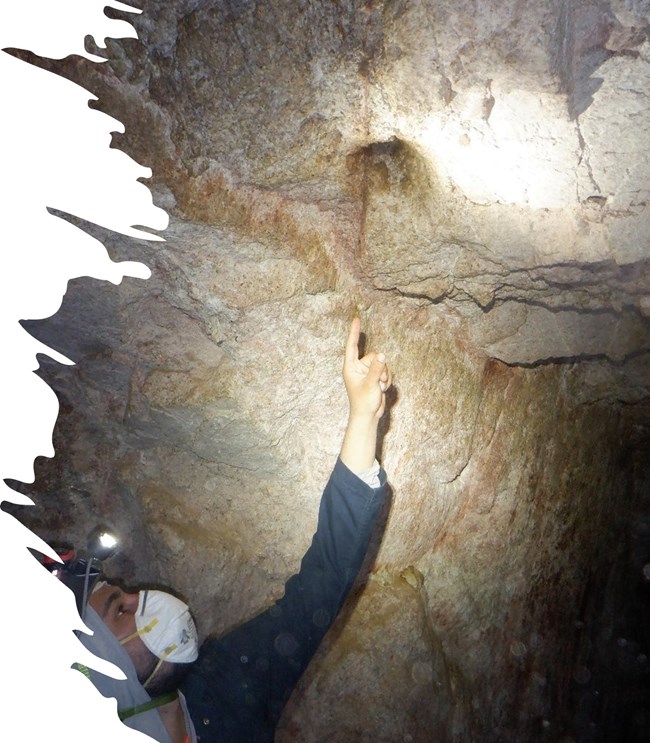 a person with a headlamp and N95 mask points to a hole in the ceiling of a cave