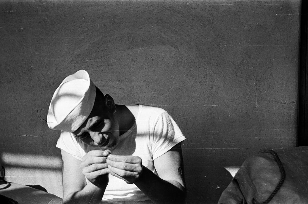 Man in sailor hat and white shirt sitting on bed threading a needle.
