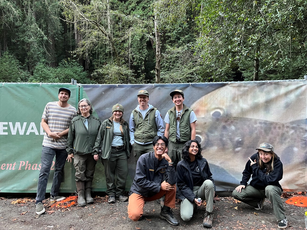 Eight park staff and interns gather for a photo in front of a banner Muir Woods featuring a photo of juvenile coho salmon.