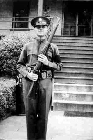 John Bishop stands in front of barracks in uniform holding a rifle