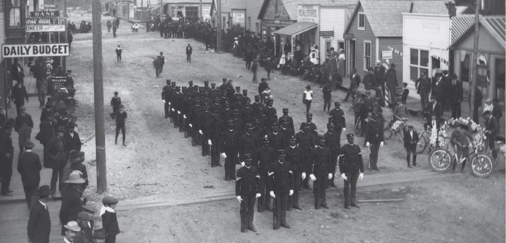 in a historic scene, black troops stand at attention on a crowded dirt road lined with buildings.