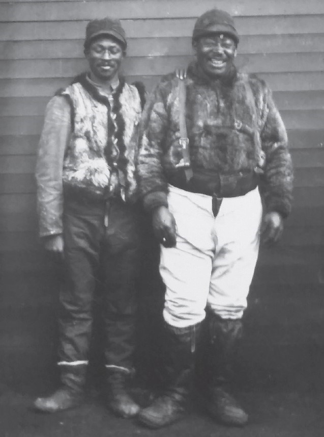 in a historic scene, two Black men smile and pose next to a building, wearing shirts of thick fur and warm boots.