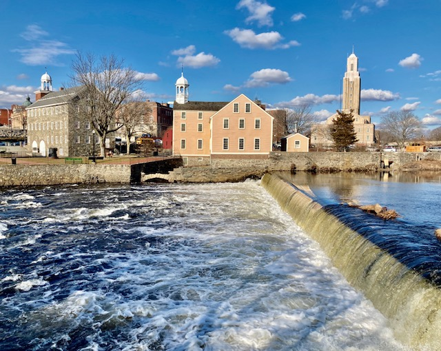 Blue river with short waterfall in foreground. Two mill buildings (one stone, one yellow wood-sided) across river. Blue skies.