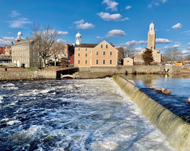 Slater Mill Blue river with short waterfall in foreground. Two mill buildings (one stone, one yellow wood-sided) across river. Blue skies.