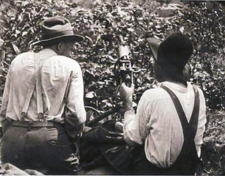 Two men in overalls and hats sit with their behind a machine gun nest amidst dead leaves and shrubs.