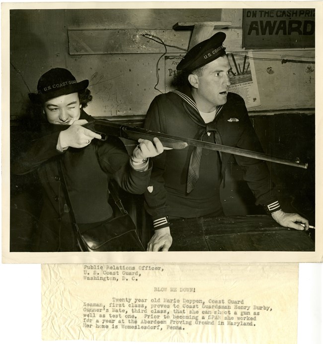 A white woman in military uniform aims a rifle while a white man in uniform stands next to her looking surprised.