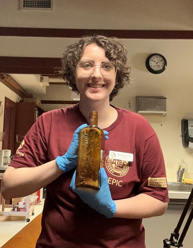 A person with short dark hair and glasses wearing an American Conservation Experience t-shirt holds a bottle made from amber colored glass.