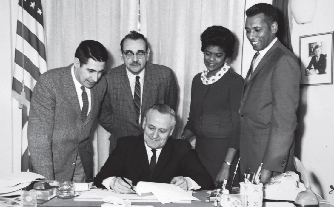 four people -- two white men and a Black man and woman -- stand behind a desk watching a man sign papers, all wearing formal dress.