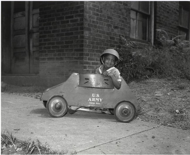Black and white photo of African American boy in pith helmet riding a toy car labeled US Army
