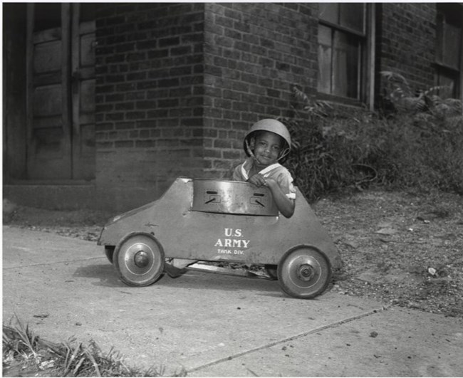Charles “Teenie” Harris; National Museum of African American History and Culture Black and white photo of African American boy in pith helmet riding a toy car labeled US Army