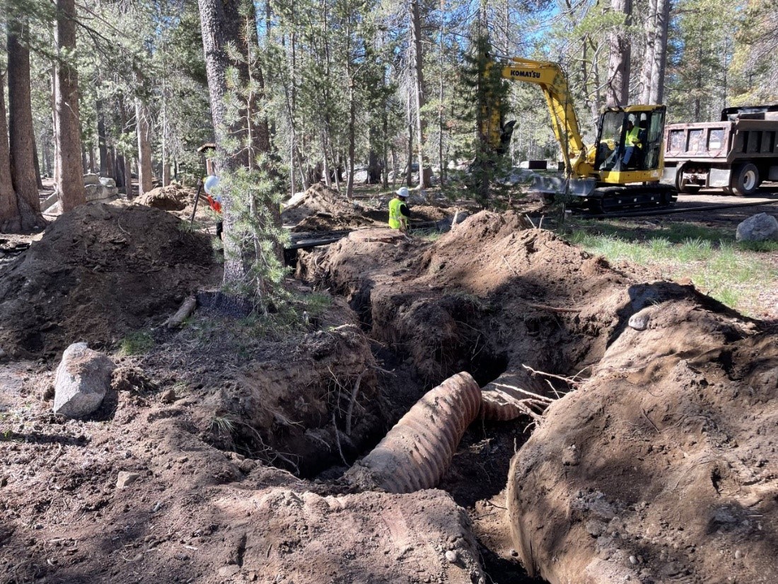 a large dirt hole exposes a large pipe. construction equipment and trees are in the background