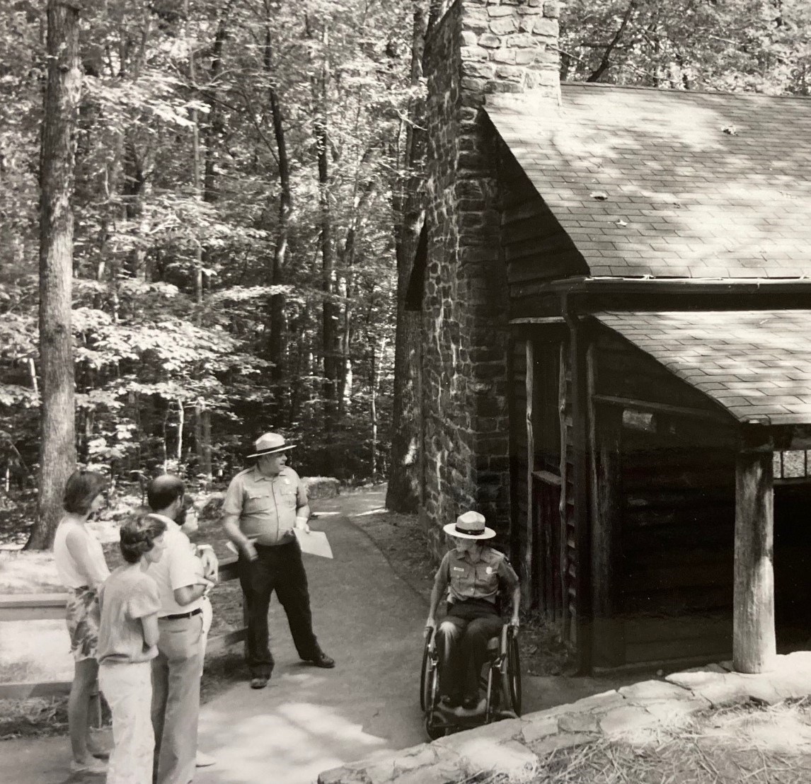 Erin Broadbent in a wheelchair and a male ranger with visitors outside a cabin.