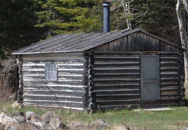 Corner view of a chinked log shack with a metal chimney in the center of the roof.
