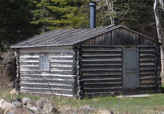 bunkhouse Corner view of a chinked log shack with a metal chimney in the center of the roof.