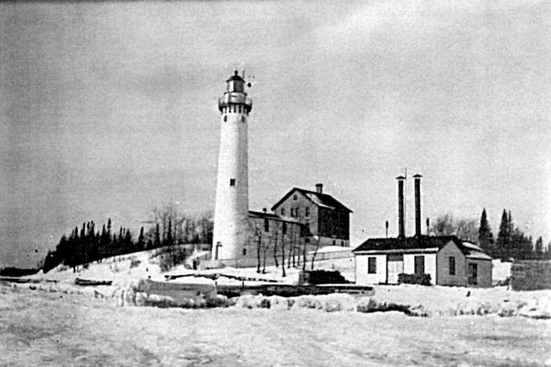 South Manitou Island Lighthouse complex A black and white image of a tall cylindrical building on a small hill with a small T-shaped building to the left.