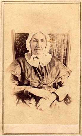 A sepia-toned photo of a woman in mid-1800s attire including a bonnet, seated in a chair.