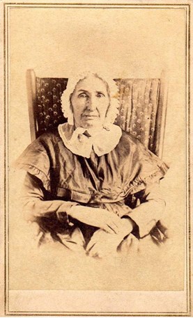 Mehitable Bradford Frazee A sepia-toned photo of a woman in mid-1800s attire including a bonnet, seated in a chair.