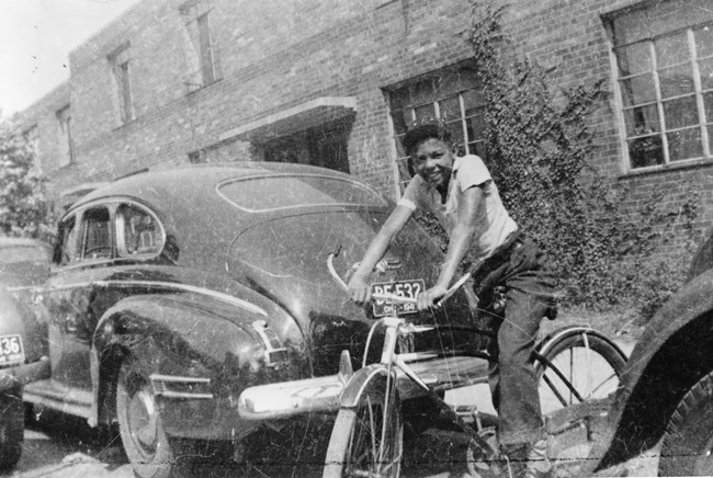 A smiling Black teenage boy sits on a bicycle between cars parked outside a brick building.