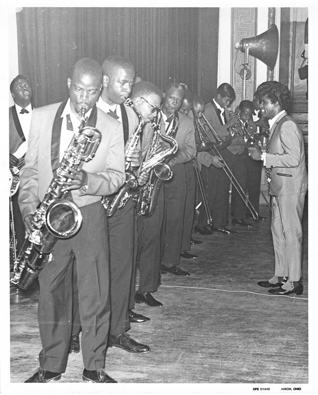 James Brown, right, faces a line of 10 Black musicians in formalwear blowing into brass instruments.