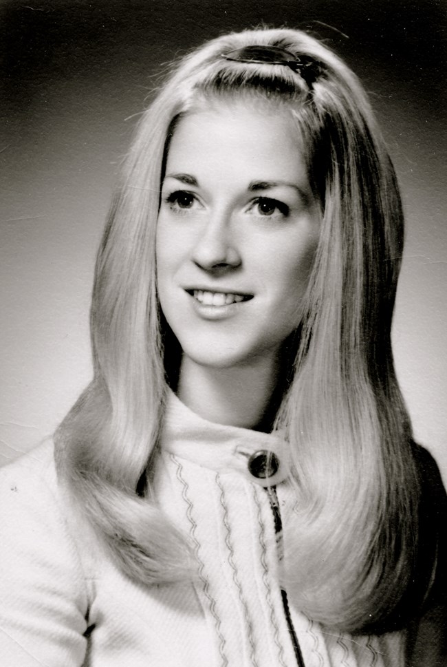 Black and white photo of a young woman posing. Her blond hair is secured with a center barrette and her pale top has vertical lines of embroidery.