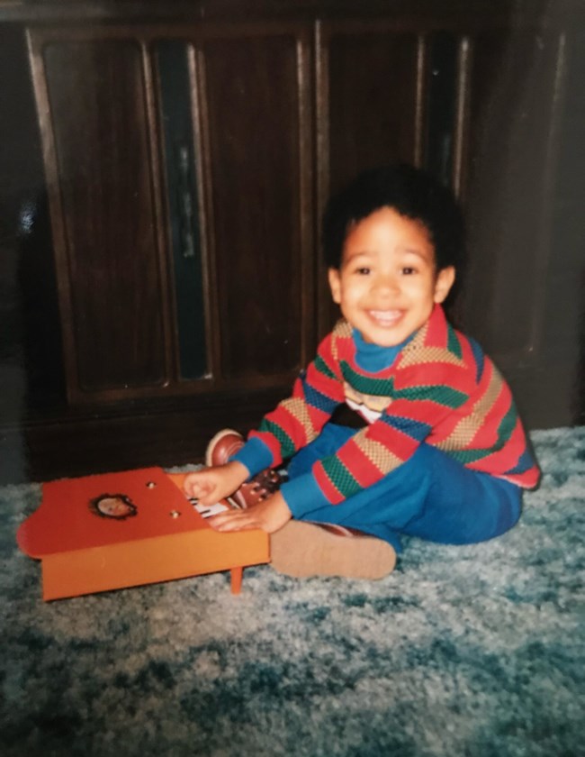 A young Black child in a striped sweater and blue pants sits smiling with his hands on a toy piano.