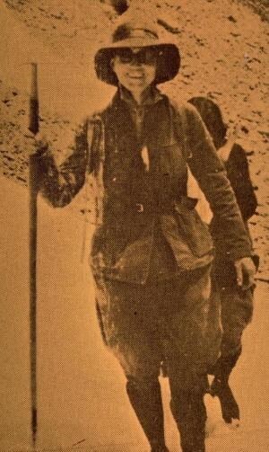 Elizabeth Burnell wearing breeches, boots, shirt, coat, had, and sunglasses uses a walking stick as she hikes ahead of another person.