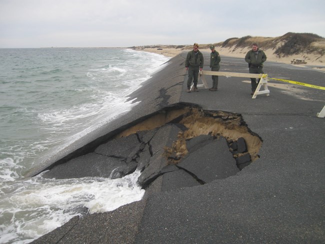 Three park staff look at a section of parking lot that has crumbled into the ocean