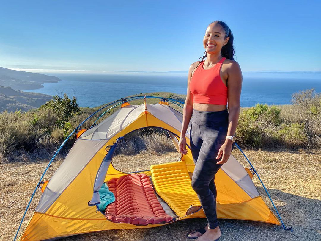 a smiling woman stands next to a tent with the ocean in the background