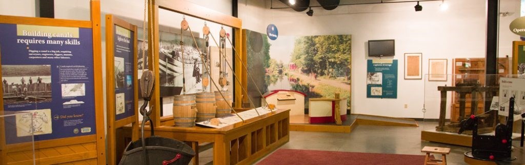 National Canal Museum Interior of museum gallery with various artifacts and displays including pulley systems. Museum text panel at left reads "Building canals requires many skills"