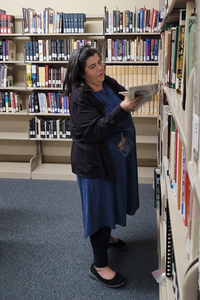 Woman standing in a library smiling at a book she has open.