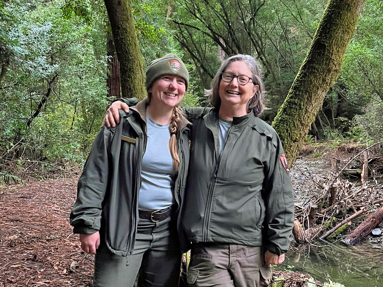 Two women wearing National Park Service jackets smile as they stand side-by-side in a forest beside the clear, calm water of Redwood Creek.