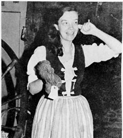 Cathy Ingram in a period costume of skirt, blouse, and vest signs with one hand and holds a skein of yarn in the other.