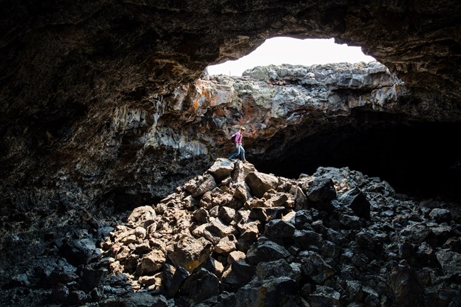 A visitor stands inside a lava tube underneath a large opening to the sky.