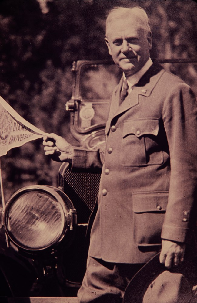 Stephen Mather in uniform standing next to car holding a pennant