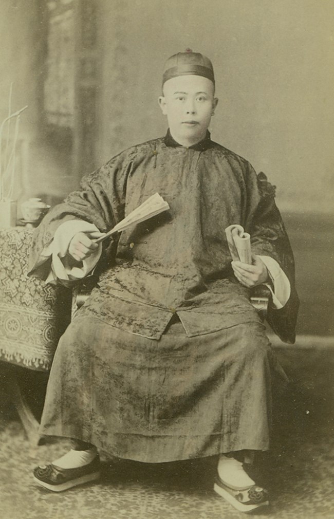 Black and white photo of a Chinese man in traditional clothing sitting in a portrait studio.