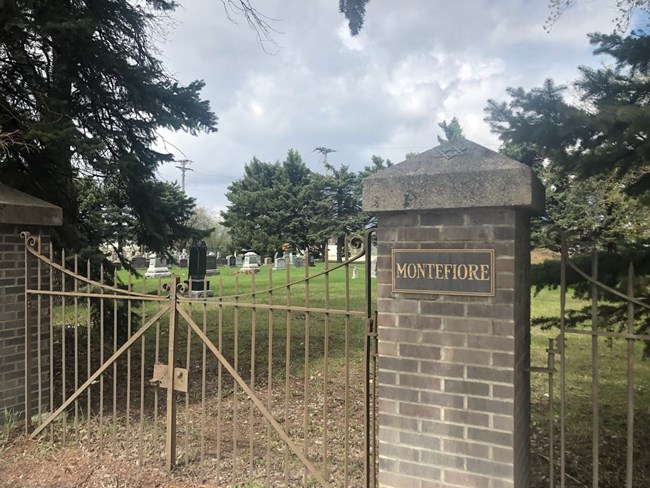 Montefiore Cemetery gate A metal gate to a cemetery. A brick post with concrete cap is on each side of the gate. A plaque with "MONTEFIORE" is on the near post, and a small Star of David is on the post's concrete cap.