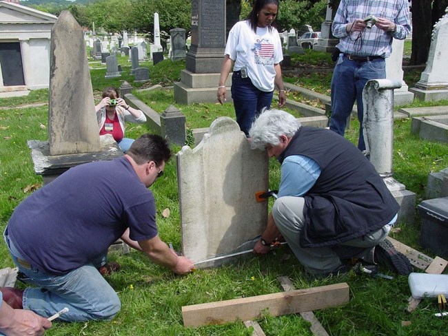 Two people carefully reset a stone grave marker and two others observe and document the repair.