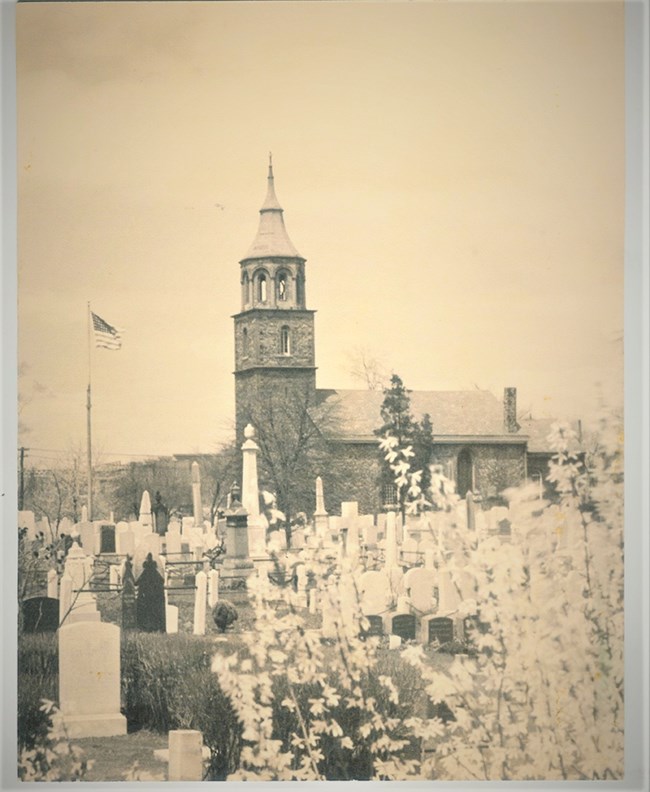 Cemetery, with gravestones of various sizes & shapes, church and flag pole visible in distance