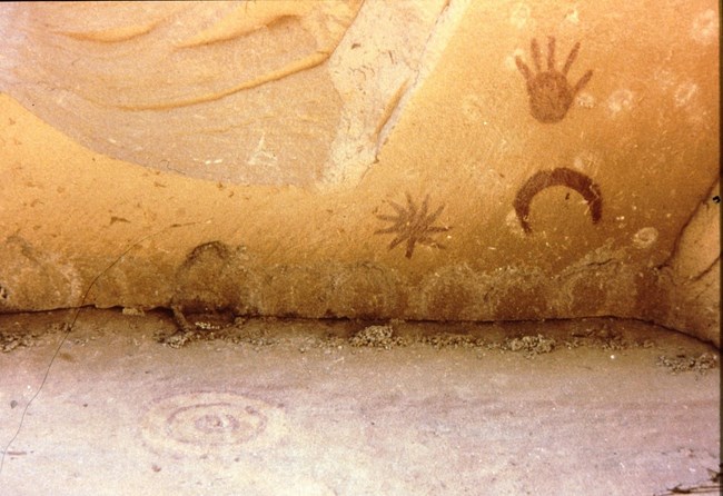 The "Supernova Pictograph" at Chaco Culture National Historical Park A red set of pictographs of a star, moon, and hand on a brown wall.