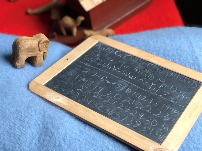 A chalkboard with letters and numbers in a child's handwriting. The chalkboard is lying on a blue felt blanket. Nearby are carved wooden animal figurines and a wooden model of Noah's ark.