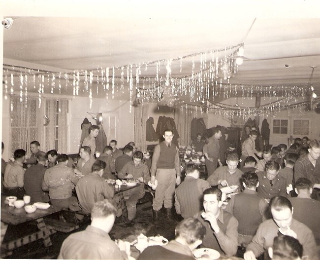 Black and white photo of uniformed men sitting at tables in a mess hall with festive decorations hanging from the ceiling.