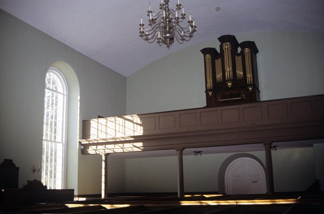 scene of the interior of a church, with balcony and an organ, and tall walled boxed pews