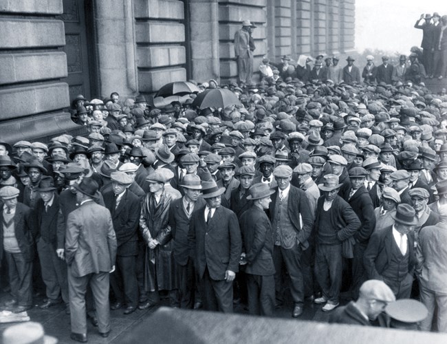 Black and white photo of a crowd of men in front of a stone facade; the men mostly wear jackets and ties and all wear hats.