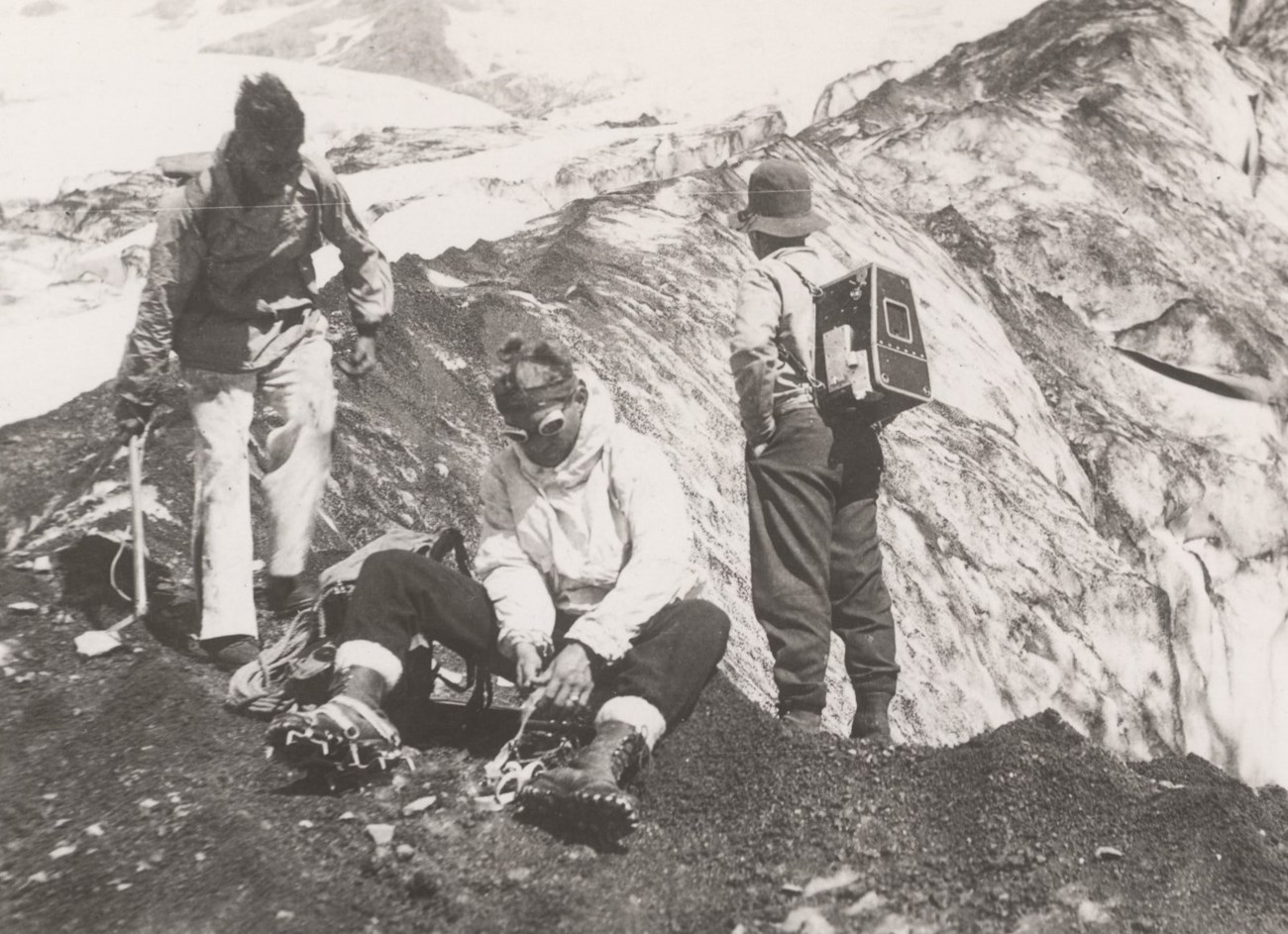 A man sits putting on ice crampons. A person with a camera case on the back stands by as another approaches the seated man.