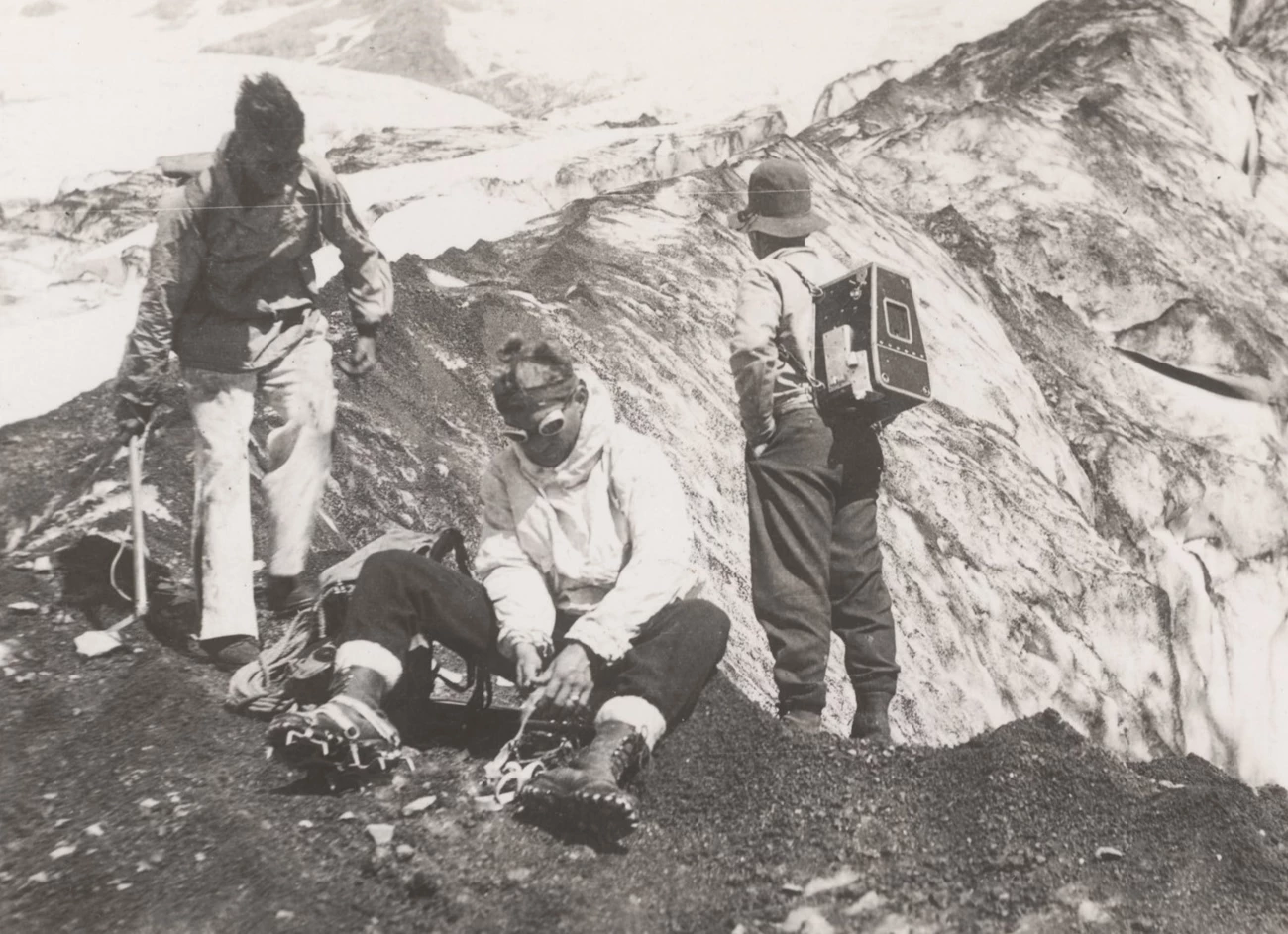 Climbers at Nisqually Glacier. A man sits putting on ice crampons. A person with a camera case on the back stands by as another approaches the seated man.