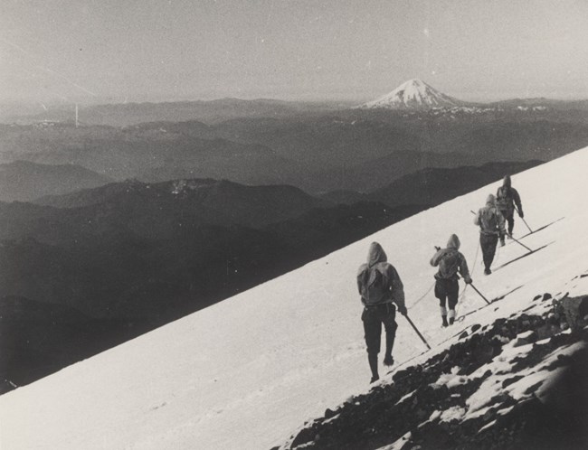 Three mountain climbers walk along a snowy mountain slope. Tall mountain in the background.